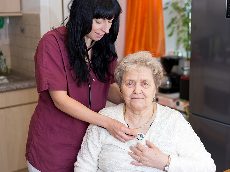 Pflegekraft hört eine Seniorin mit dem Stethoskop ab und kontrolliert ihre Gesundheit bei einem Hausbesuch einfühlsam.